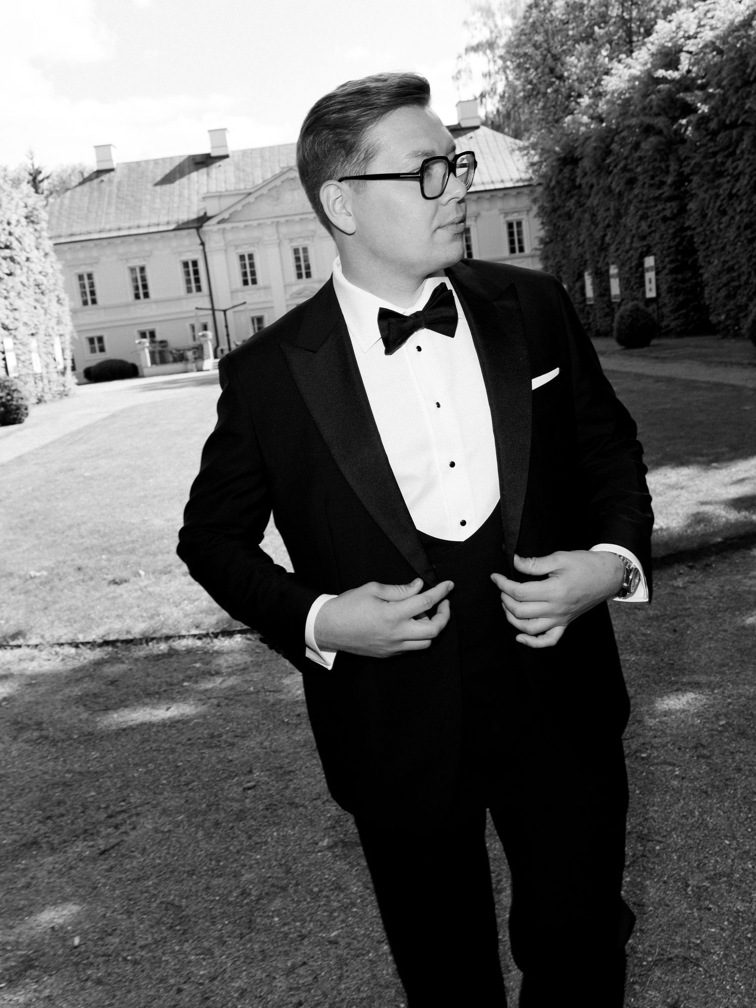 Man in black tuxedo with bow tie and glasses posing outdoors near a historic building in a black and white photo.