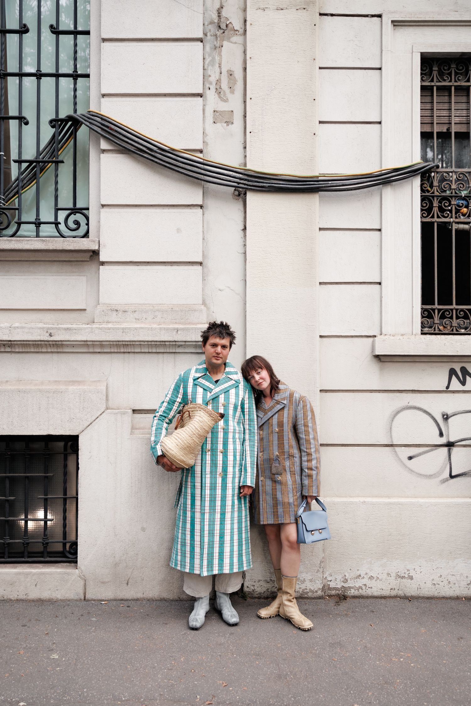 Two people in stylish outerwear pose against a white building wall in an urban setting.