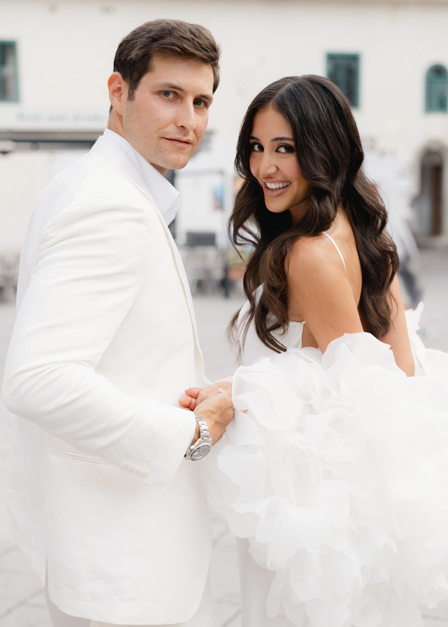 Elegant couple in matching white wedding attire posing together, bride in ruffled gown and groom in white tuxedo jacket.
