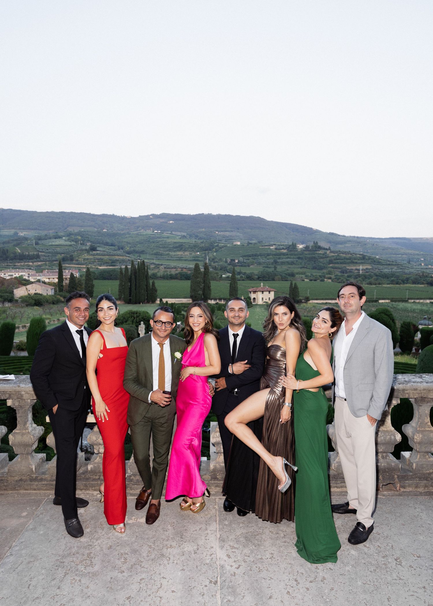 Elegantly dressed group of eight friends posing together at a scenic Tuscan villa with rolling green hills and cypress trees.