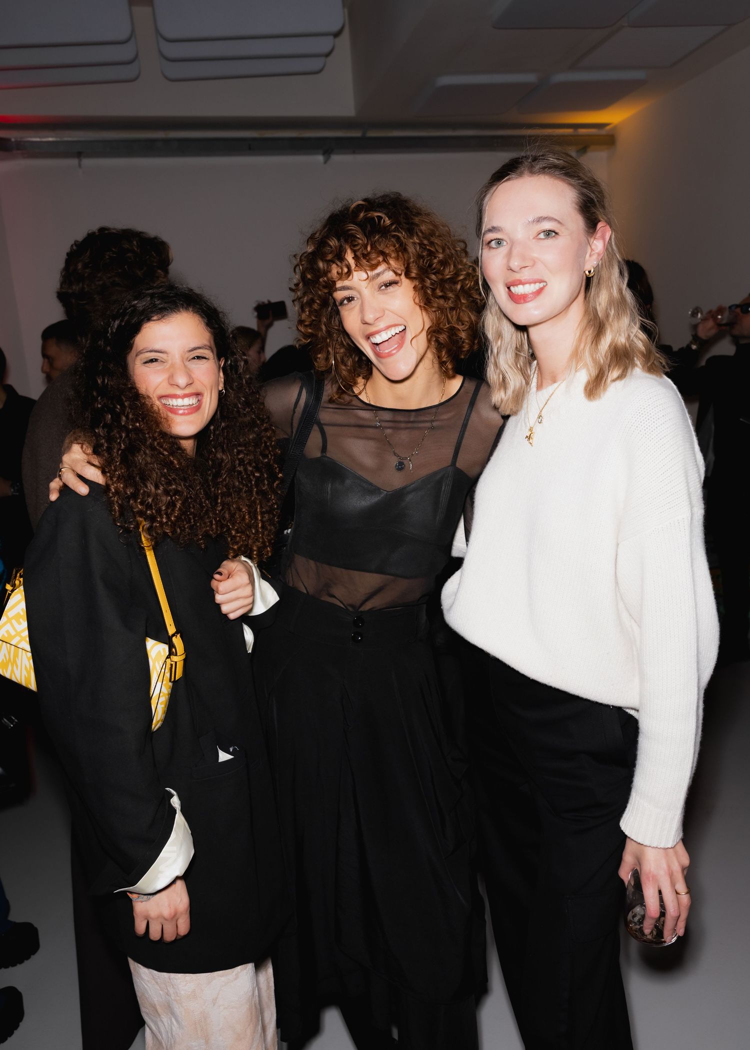 Three friends smile together at a social event wearing contrasting black and white outfits.