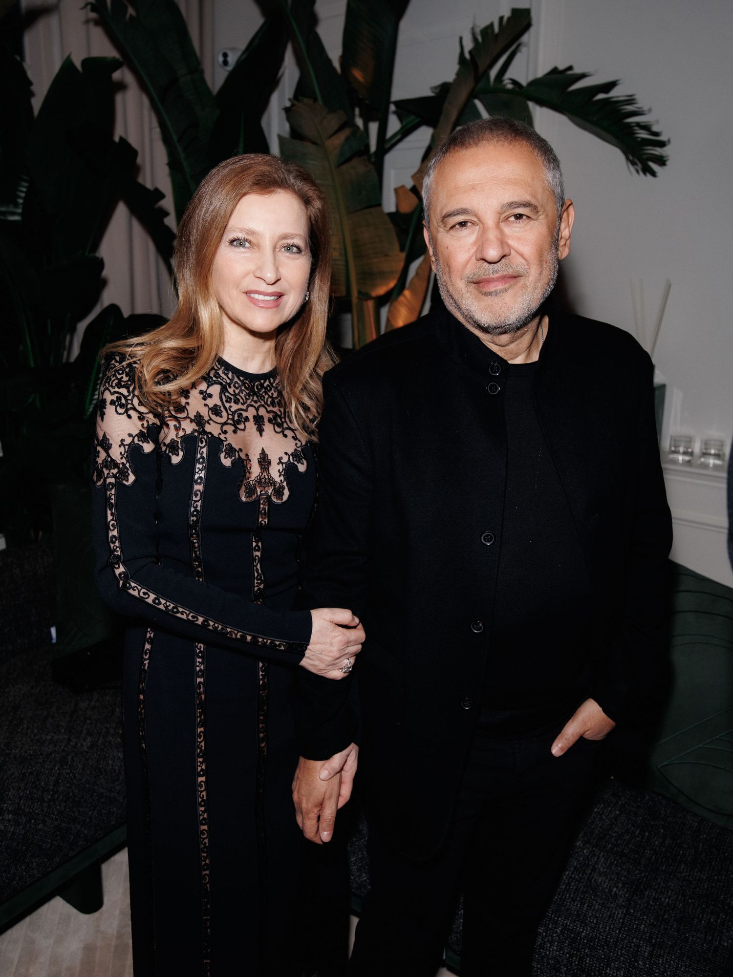 A couple wearing elegant black evening attire pose together at a formal event with tropical plants in the background.