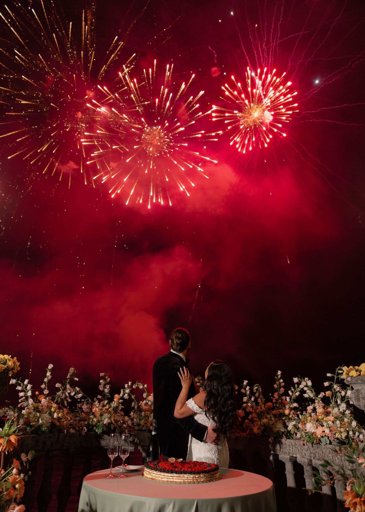Spectacular red fireworks illuminate the night sky over a wedding celebration with guests watching from below.