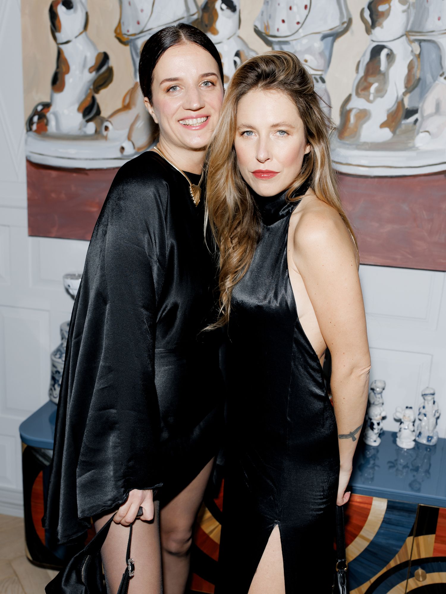 Two women in black dresses pose together against a wall with decorative figures.