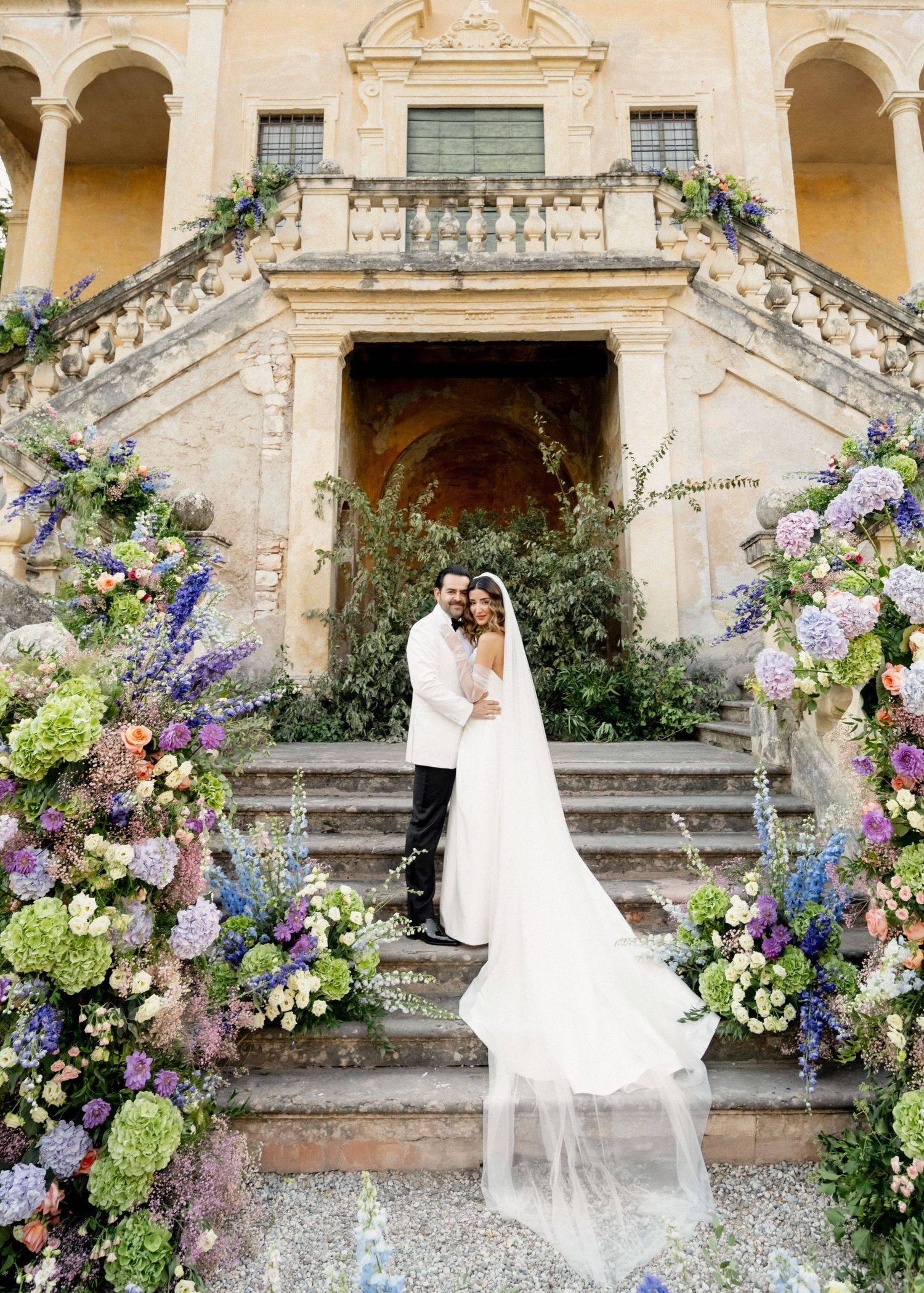 Bride and groom embrace before a grand villa staircase adorned with lush purple, blue, and green floral arrangements.