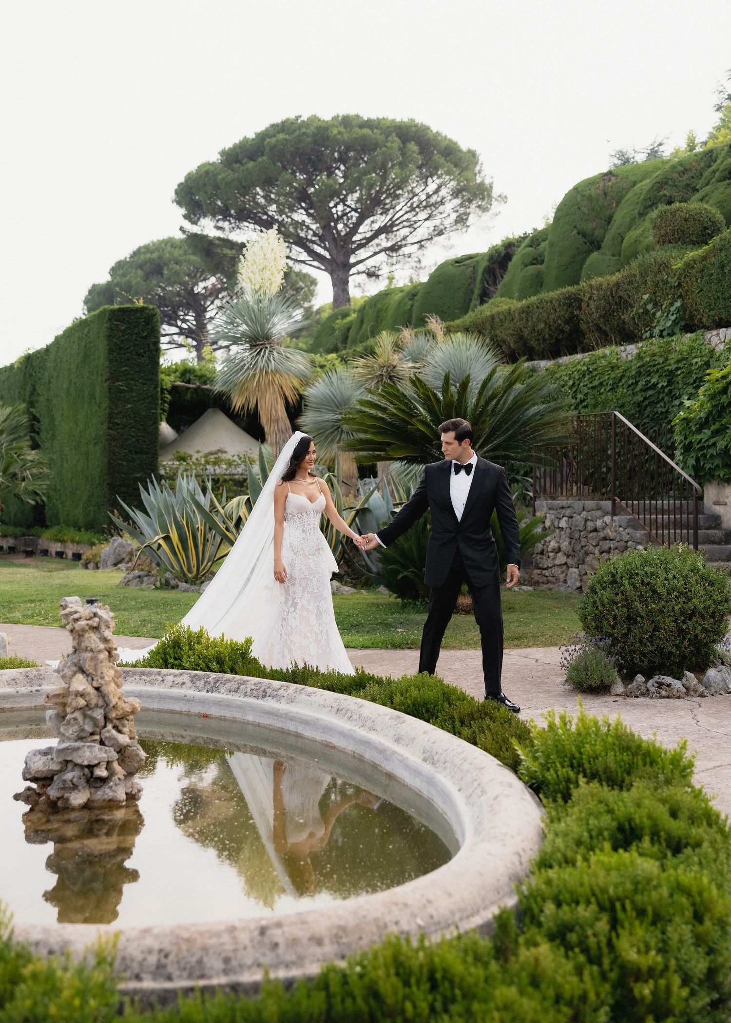 Bride and groom walk hand in hand beside a stone fountain in a lush formal garden with sculpted hedges.
