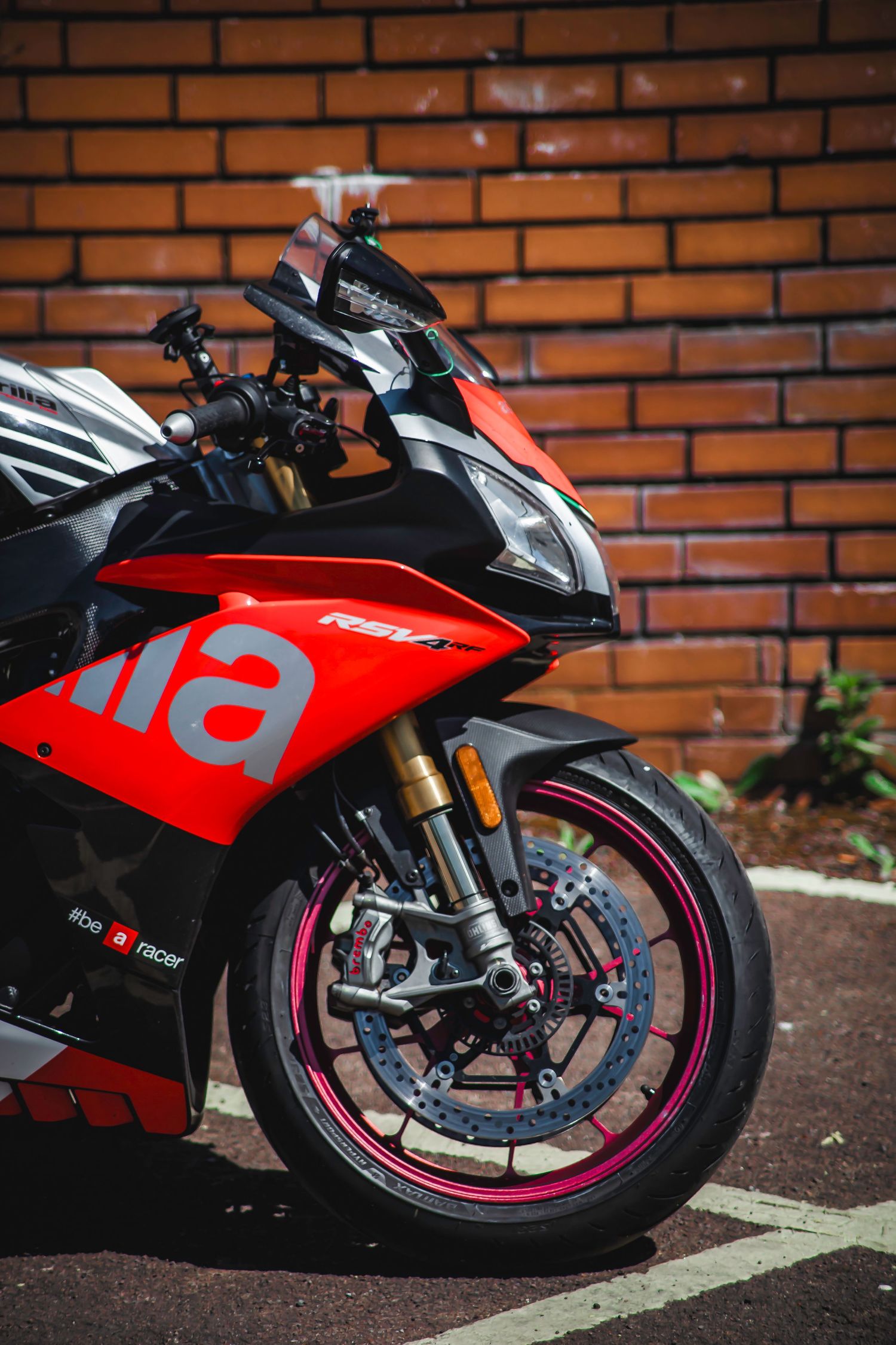 A sleek red and black Aprilia superbike parked against a brick wall, featuring gold forks and custom wheels.
