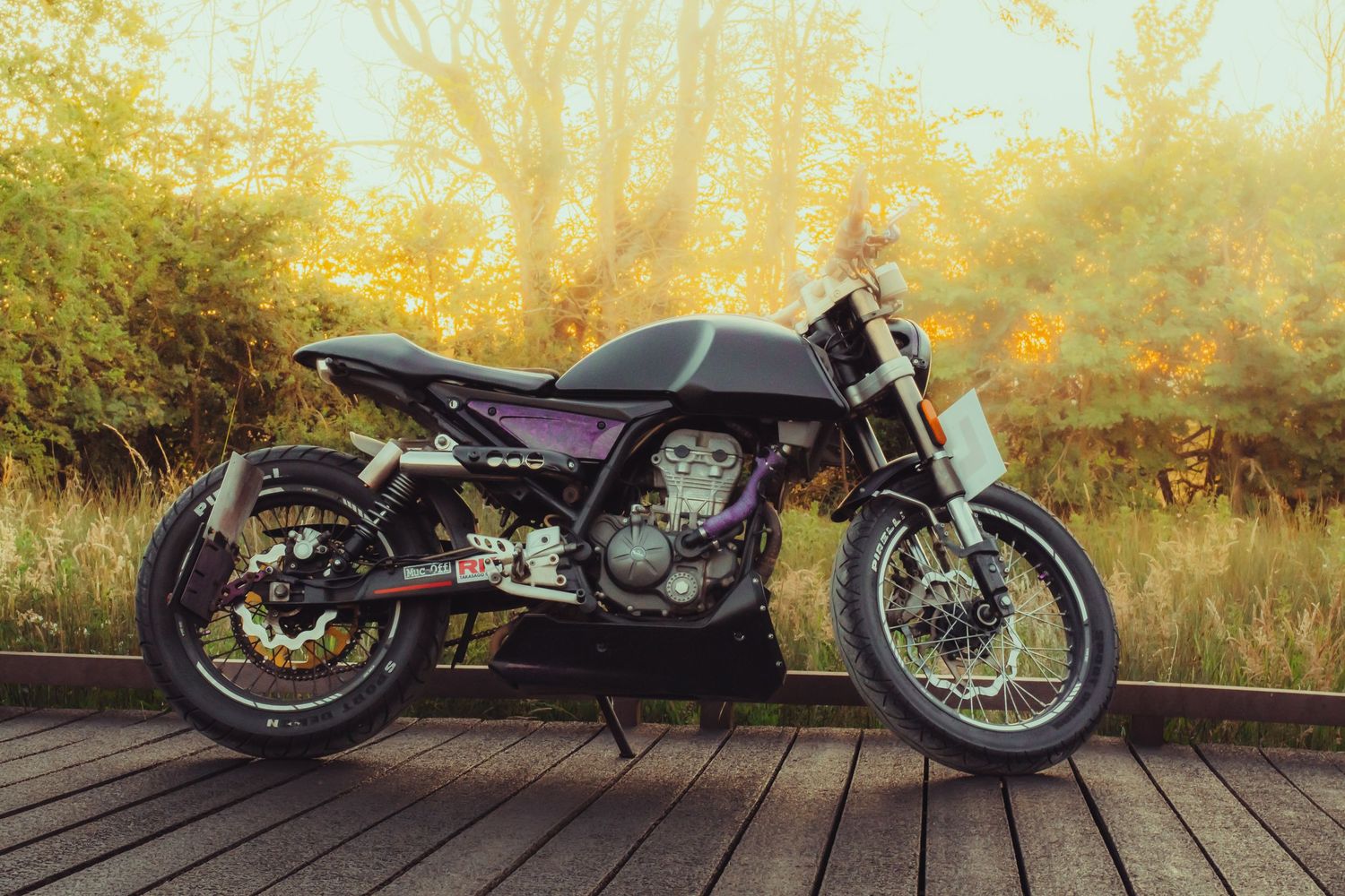 Custom cafe racer motorcycle parked on wooden deck surrounded by autumn foliage in warm golden light.