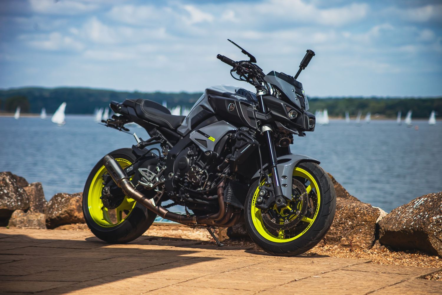 Black Yamaha MT motorcycle with neon yellow wheels parked on rocky shoreline against blue water and sky backdrop.