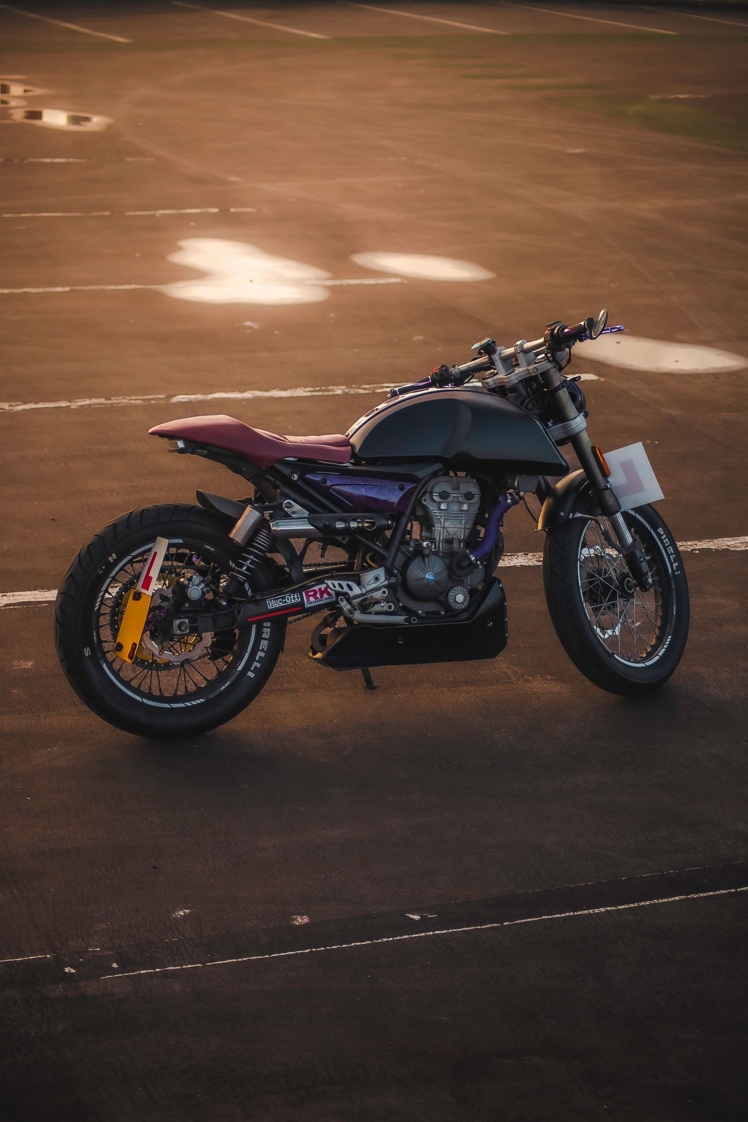 Custom cafe racer motorcycle parked on a sunlit track, featuring a sleek dark frame with purple accents and spoke wheels.