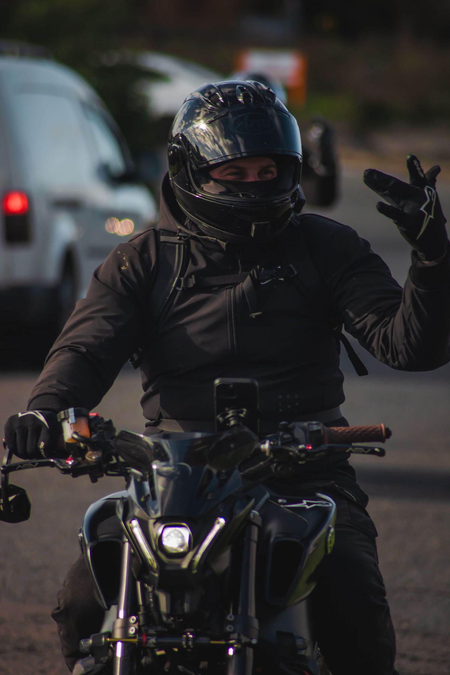A motorcyclist in black protective gear leans forward on a dark motorcycle at dusk.