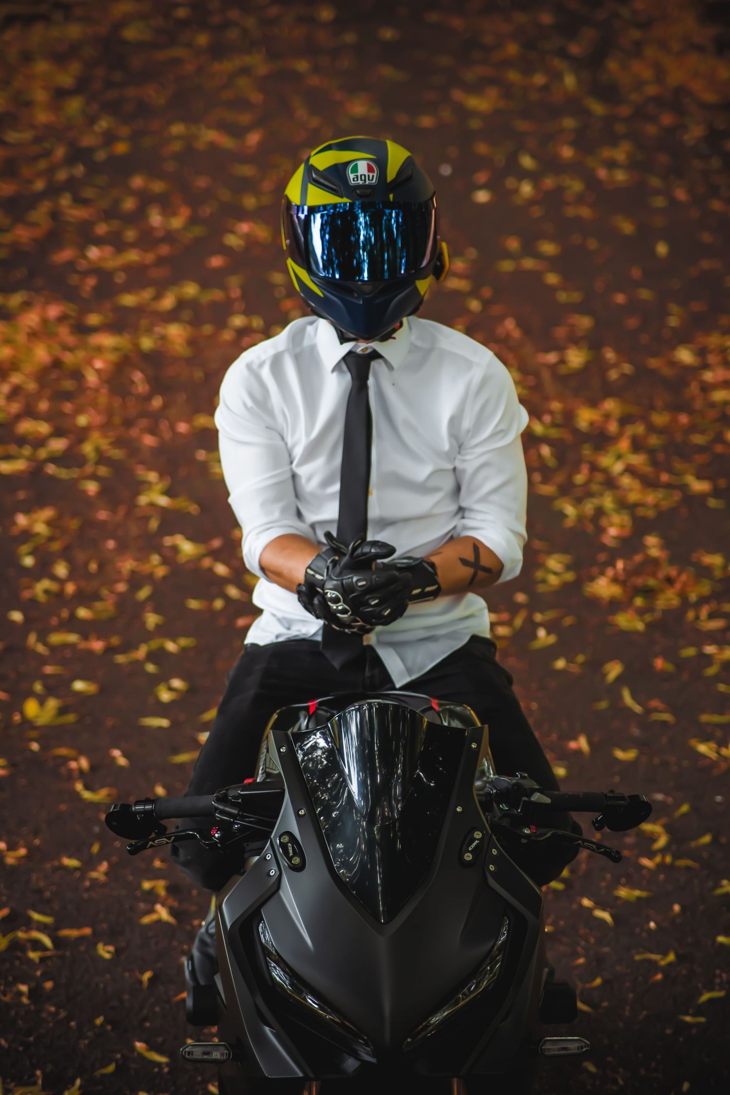 Motorcyclist in formal attire and helmet sitting on black sports bike surrounded by fall foliage.