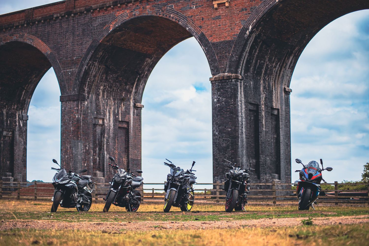 Motorcycles parked beneath a historic red brick railway viaduct against a blue sky background.