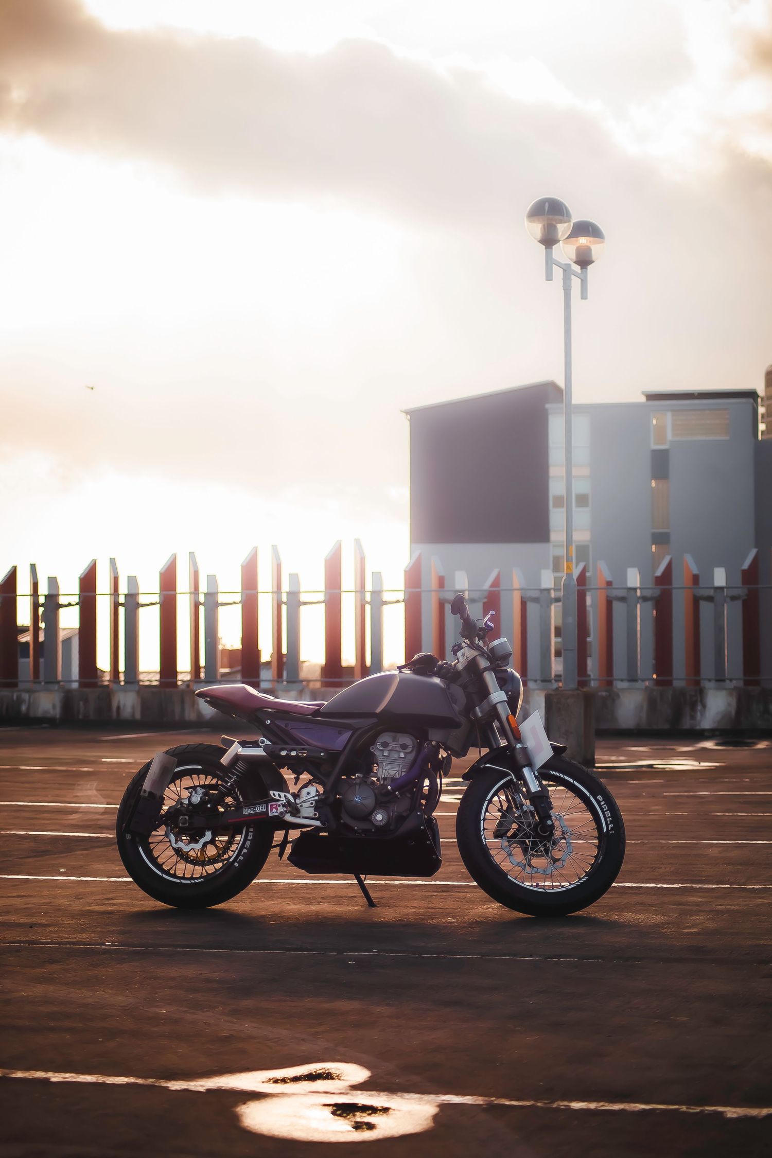 Vintage style motorcycle parked in empty parking lot at sunset with orange sky and street lamps in background.