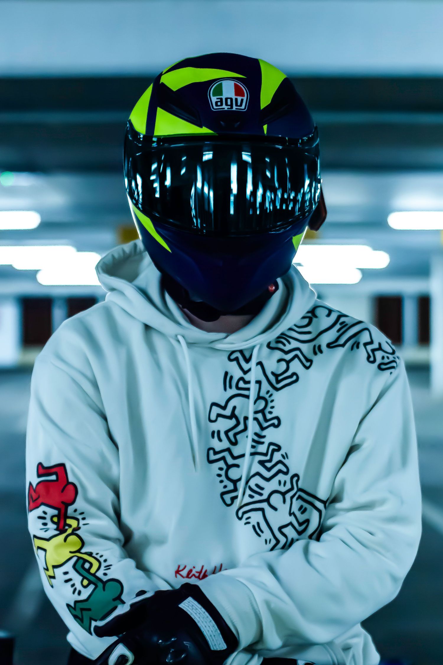 Motorcyclist wearing a white hoodie with colorful graphic designs and neon yellow helmet in a parking garage.