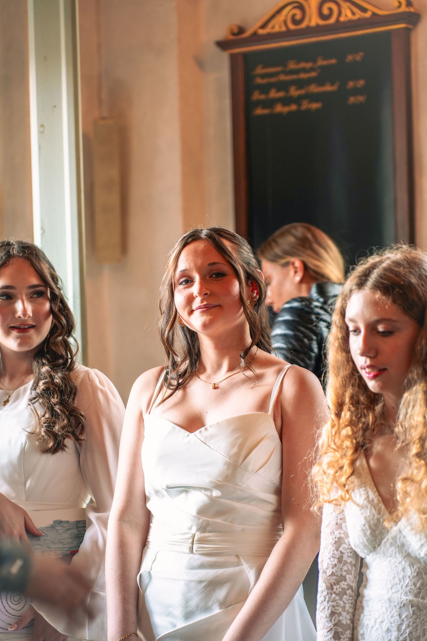 Three people wearing elegant white dresses stand together in a warmly lit indoor setting.