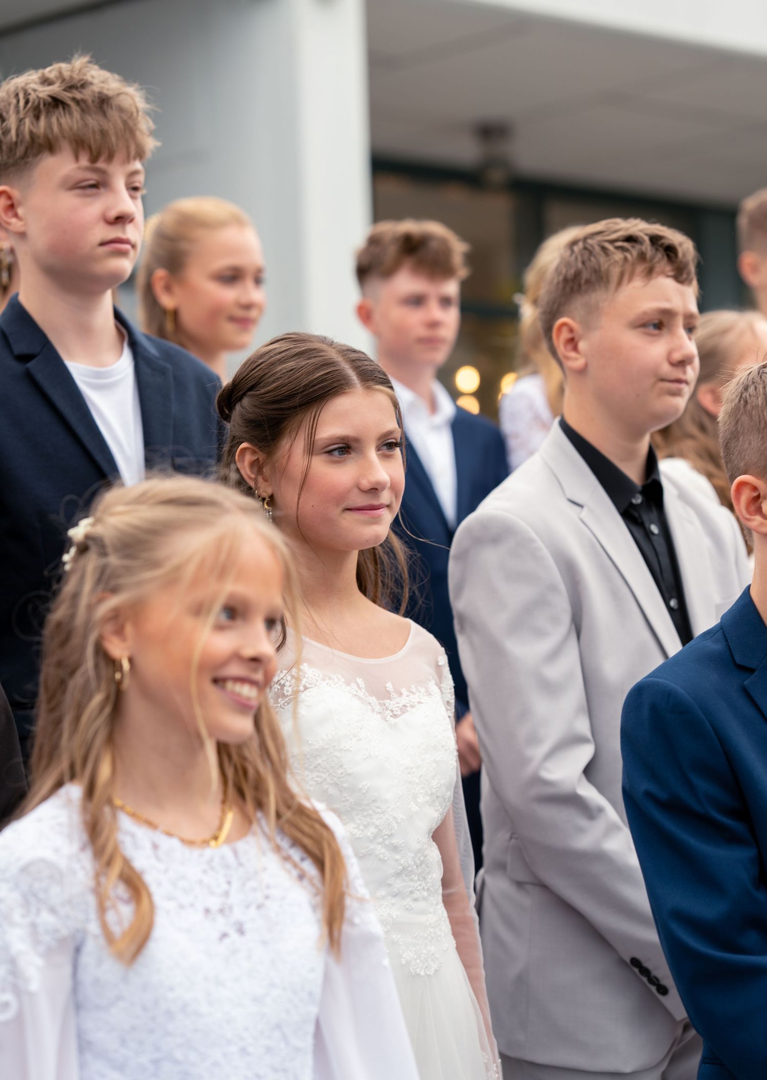 A group of young people in formal attire stand together, with some wearing suits and white dresses.