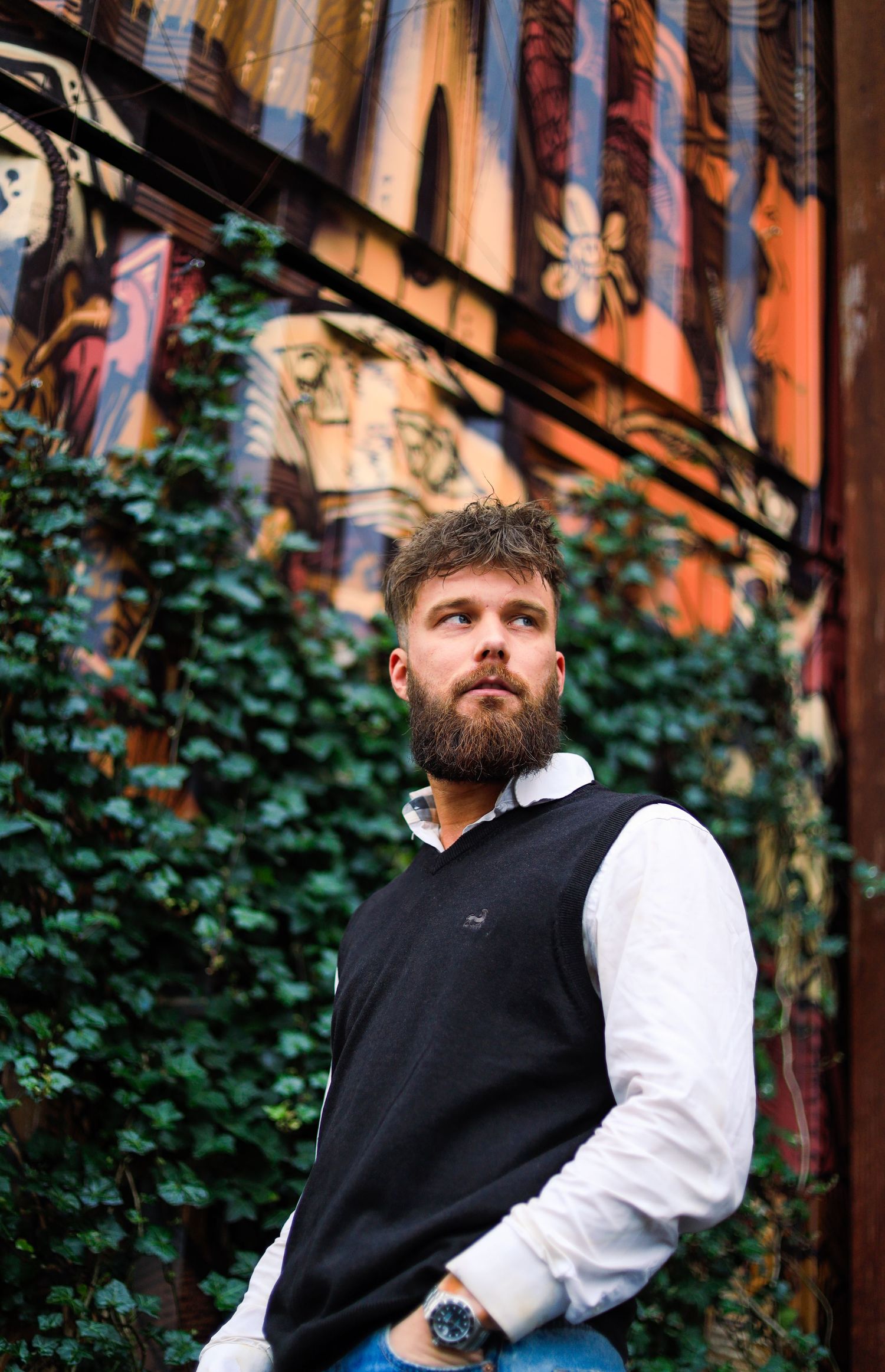 Smart casual style with black sweater vest and white shirt photographed against ivy-covered wall outdoors.