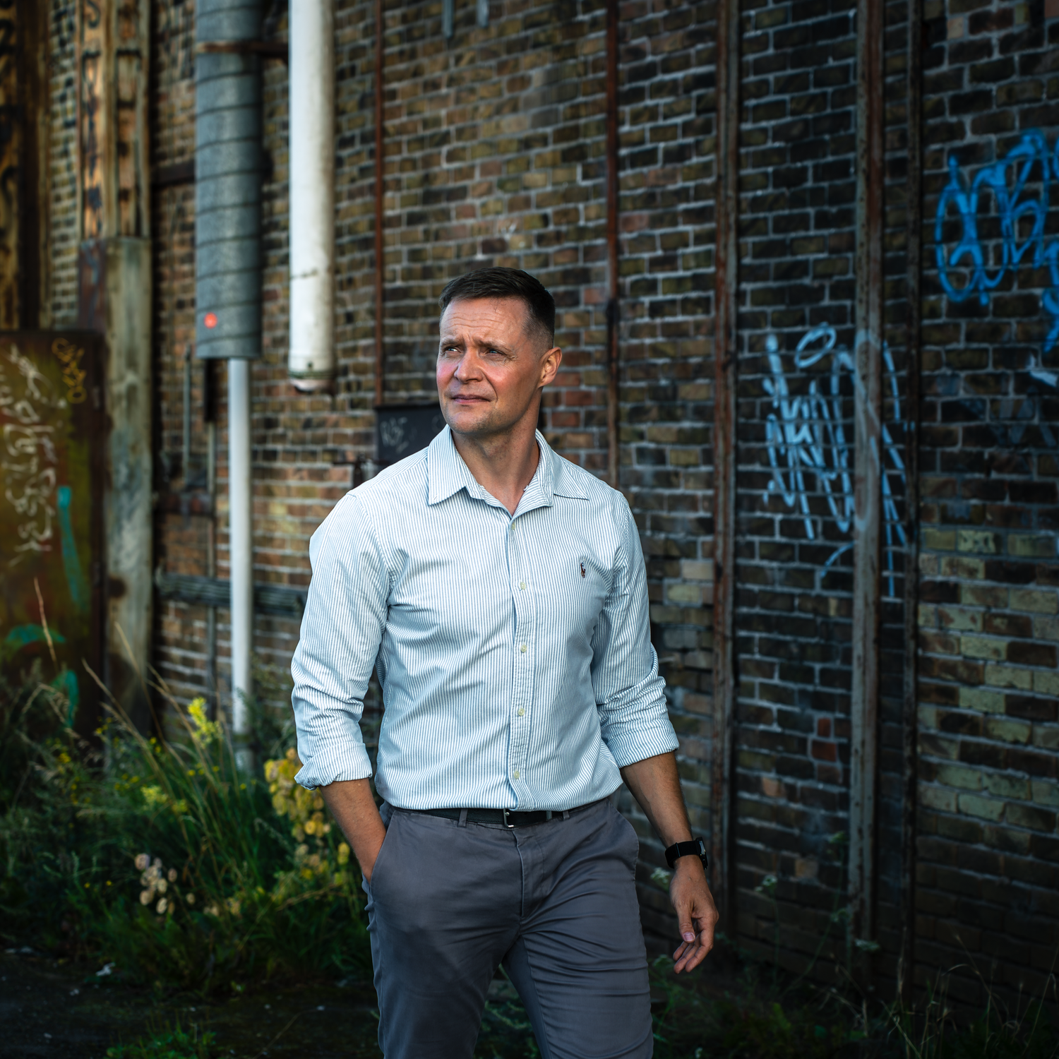 Someone in a light blue button-down shirt stands against an aged brick wall with graffiti in an urban setting.