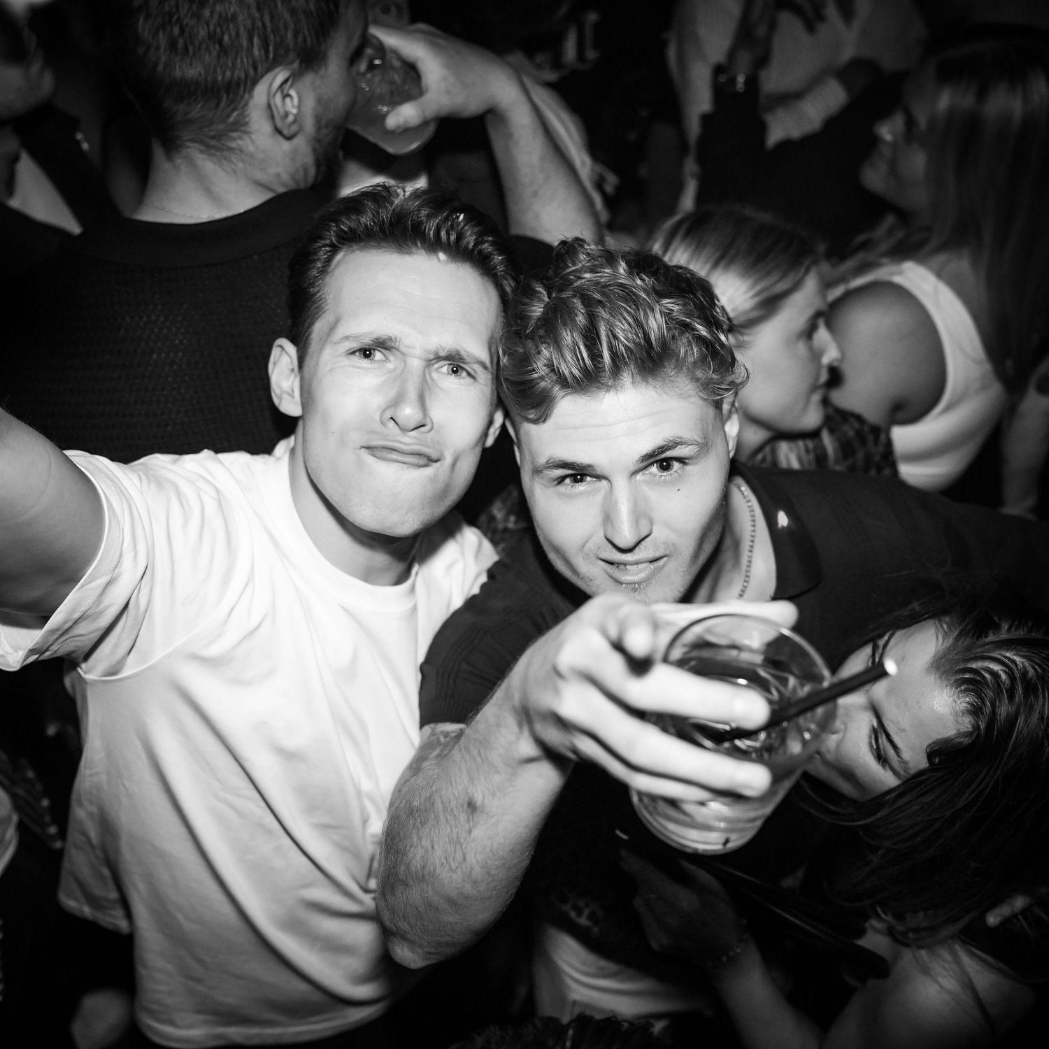 Black and white photo of two men at a nightclub, one wearing white pointing and posing for the camera.