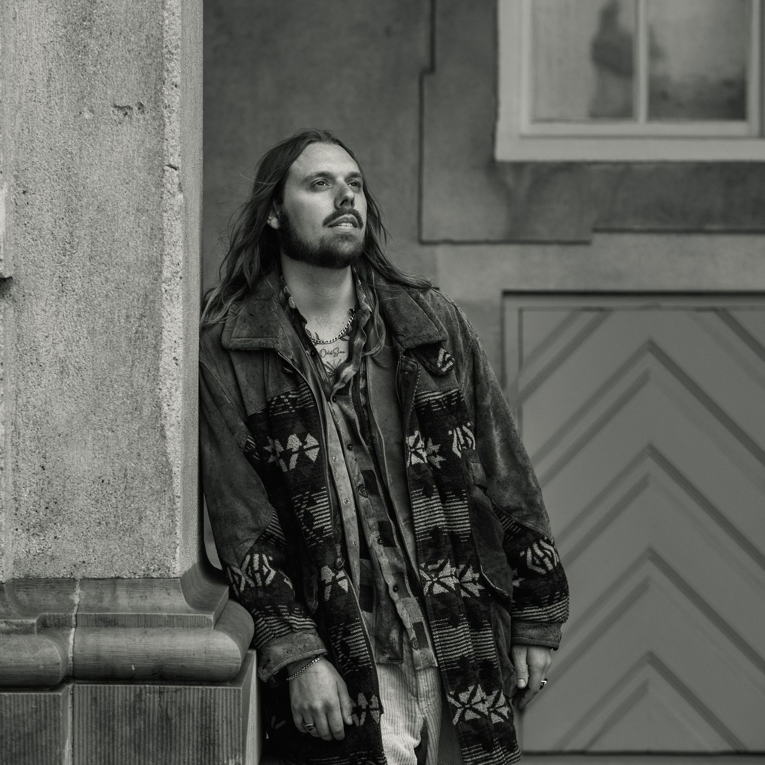 Black and white portrait of someone in embroidered bohemian style jacket gazing to the side against stone wall background.