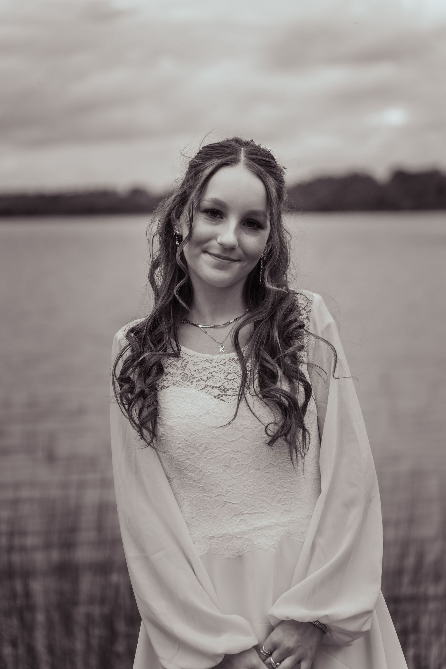 Black and white portrait of a bride in a lace wedding dress with long wavy hair standing by a lake.
