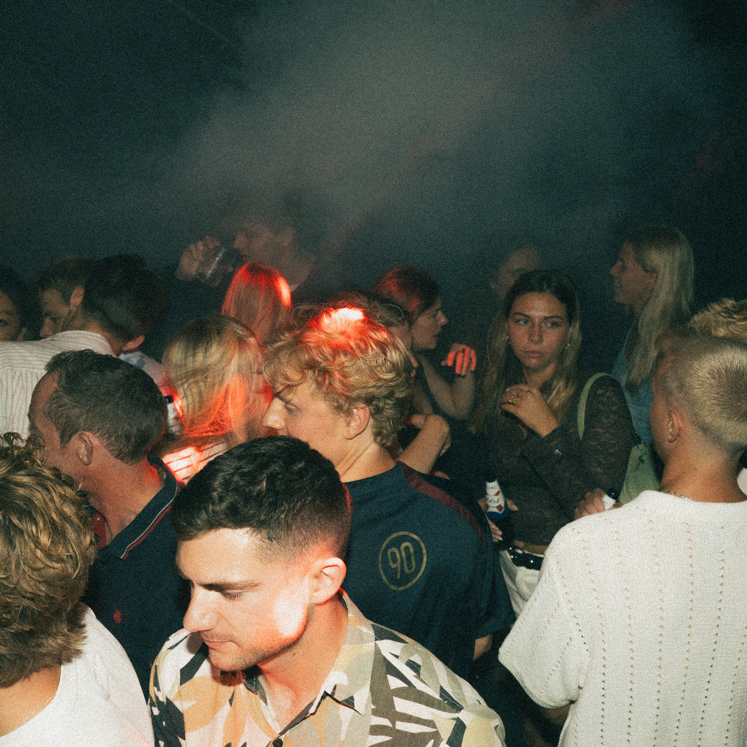Crowded dimly lit nightclub scene with people dancing and mingling in atmospheric lighting.
