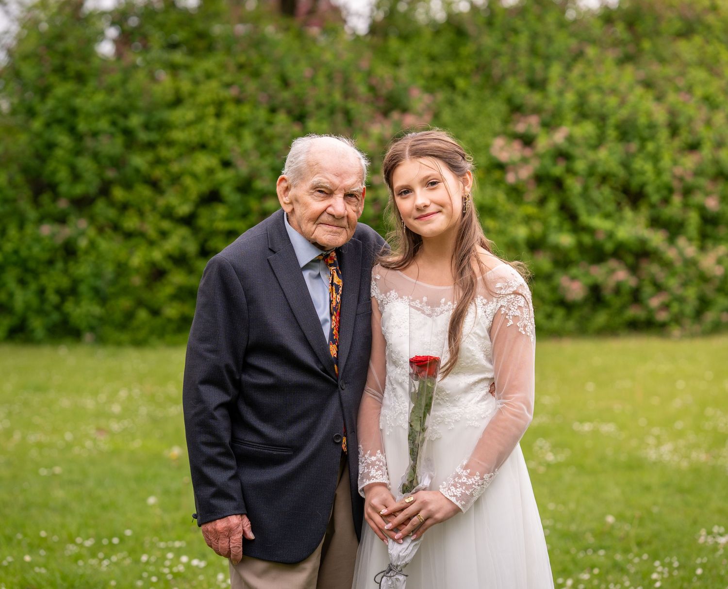 A sweet wedding portrait taken outdoors on a green lawn with flowering bushes in the background.