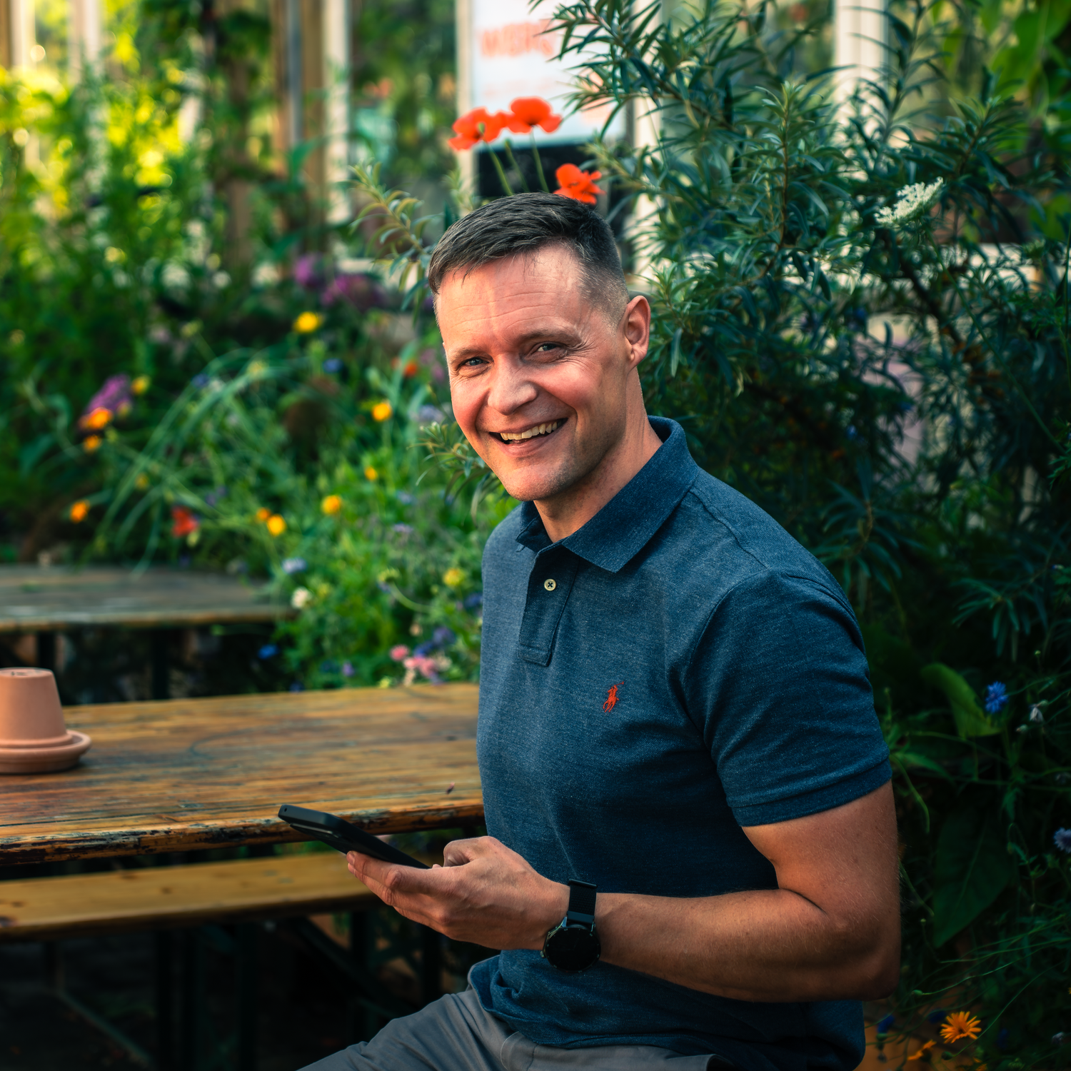 Man in navy blue polo shirt sits at outdoor wooden table in lush garden setting surrounded by greenery and flowers.