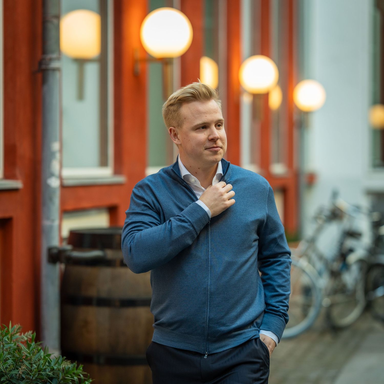Person in blue jacket adjusts collar while standing outside building with orange lights.