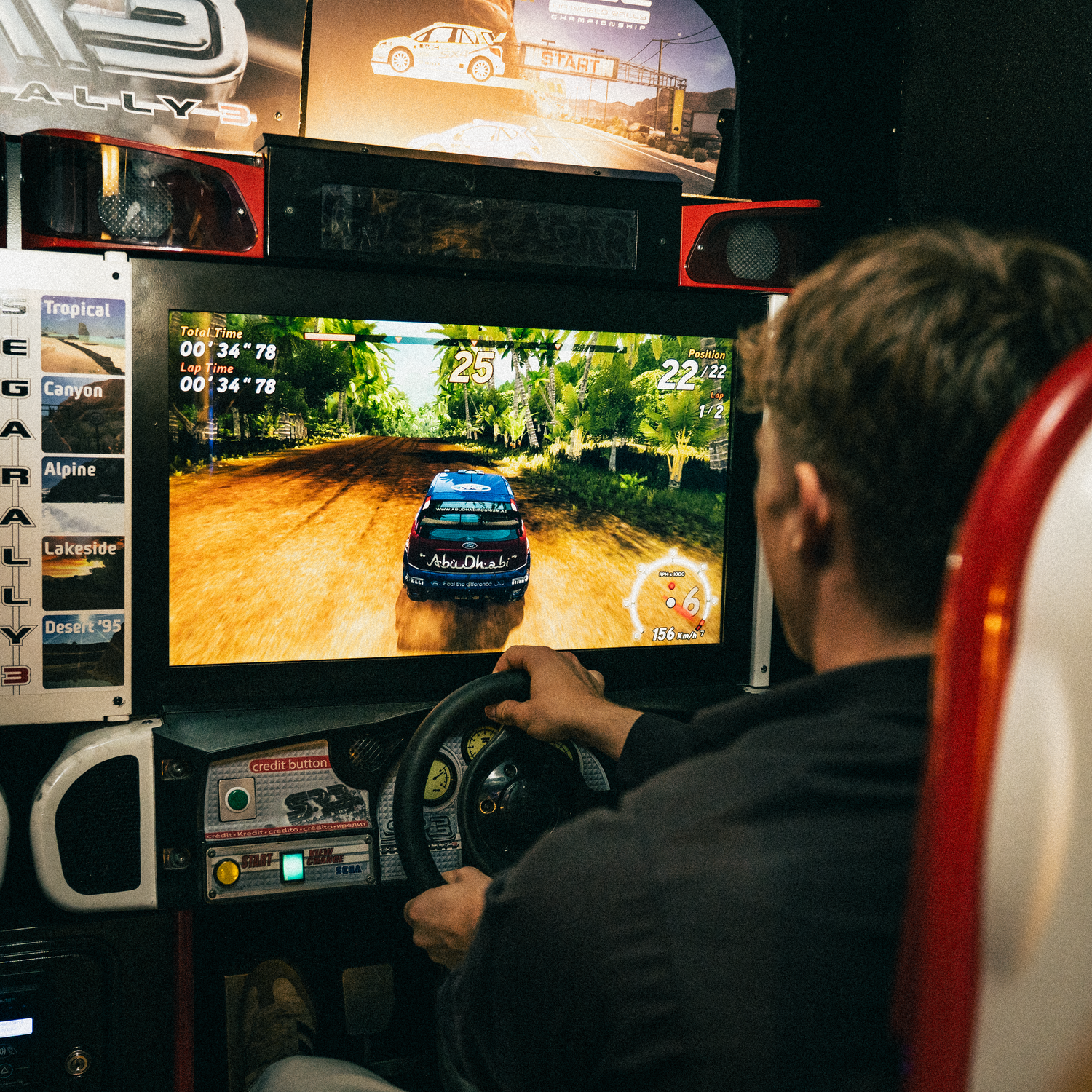 People playing virtual rally racing games side by side on large displays in a dimly lit gaming arcade setting.