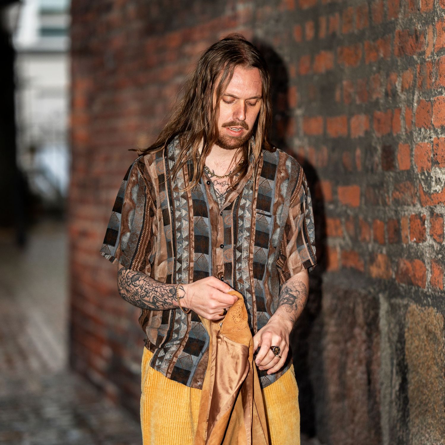 Person with long hair wearing a patterned shirt and yellow pants stands against rustic brick wall in urban alley.