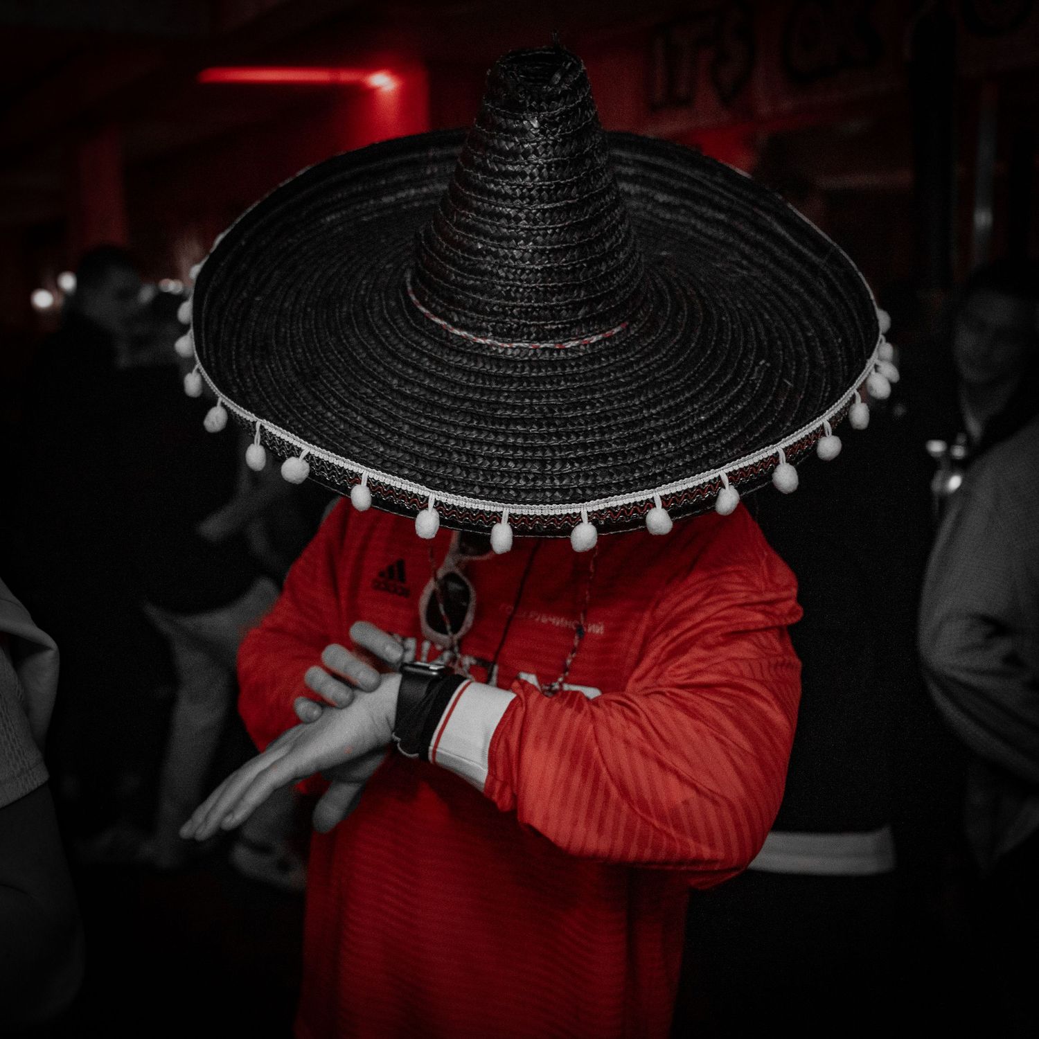 Person wearing a large black sombrero with white pom poms in a dark nightclub setting.