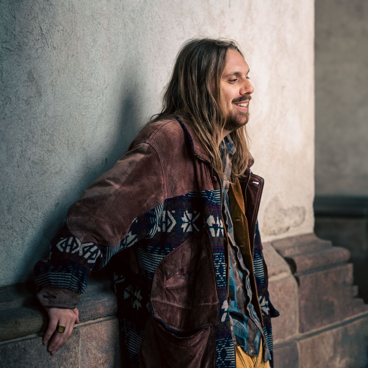 Someone with long hair and a brown suede jacket with patterned details laughs while leaning against a wall.