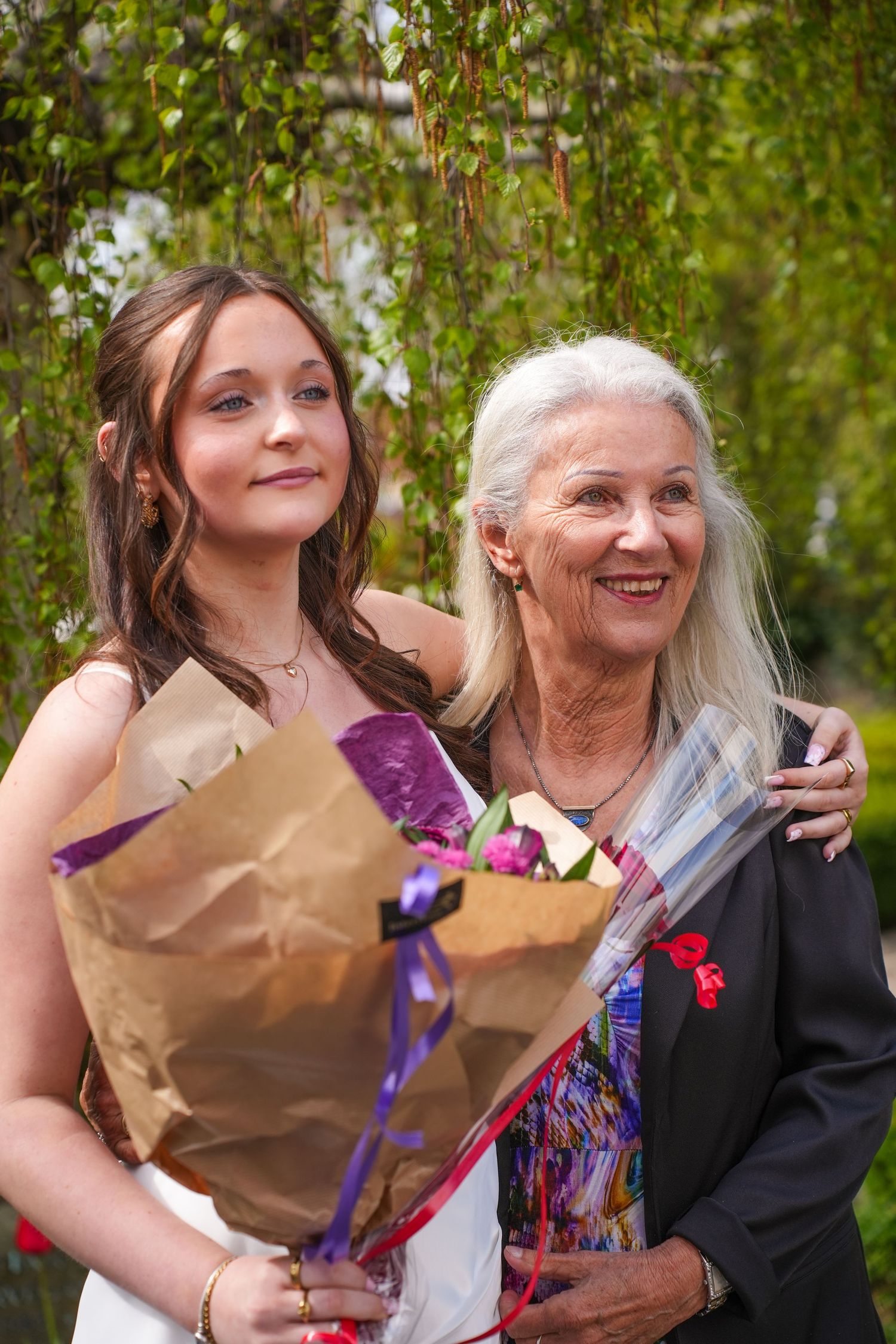 Two women standing together under a willow tree outdoors, holding flower bouquets and smiling.
