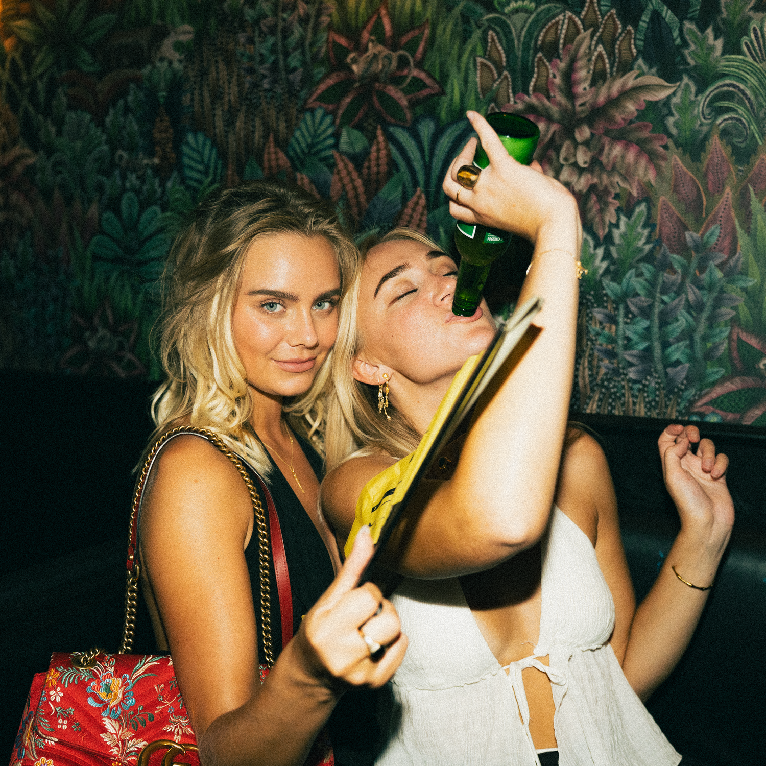 Two friends dancing and enjoying drinks at a party with tropical decor and dark lighting in background.