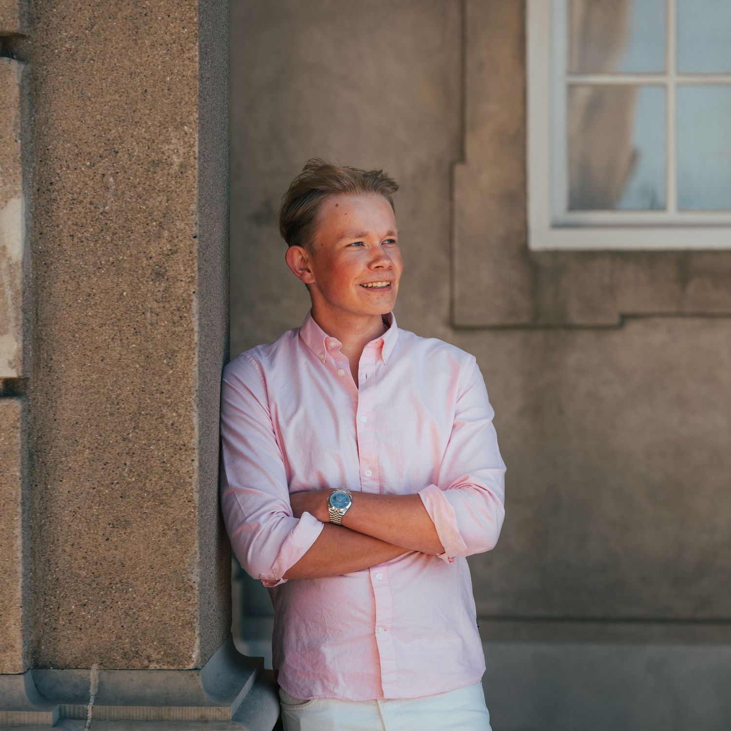 Person in pink button-down shirt standing against stone building wall with crossed arms and warm smile.