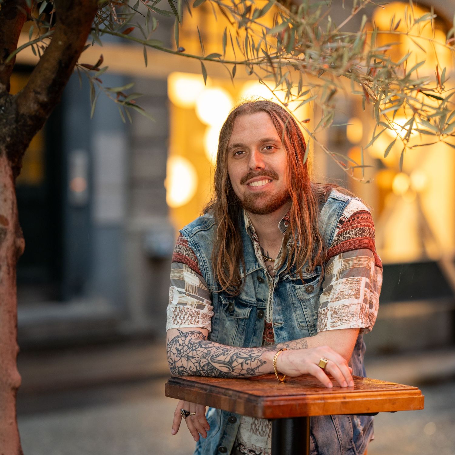 Long-haired person in denim jacket with tattoos sitting at wooden table with warm lighting and olive trees in background.