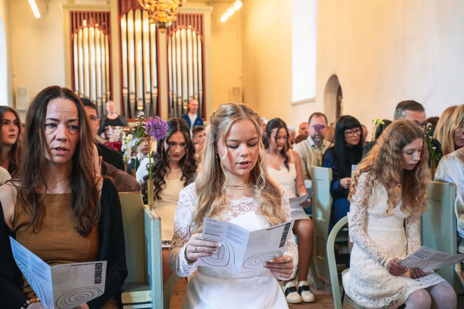 A group of attendees stand in a church with organ pipes visible, holding papers during what appears to be a service.