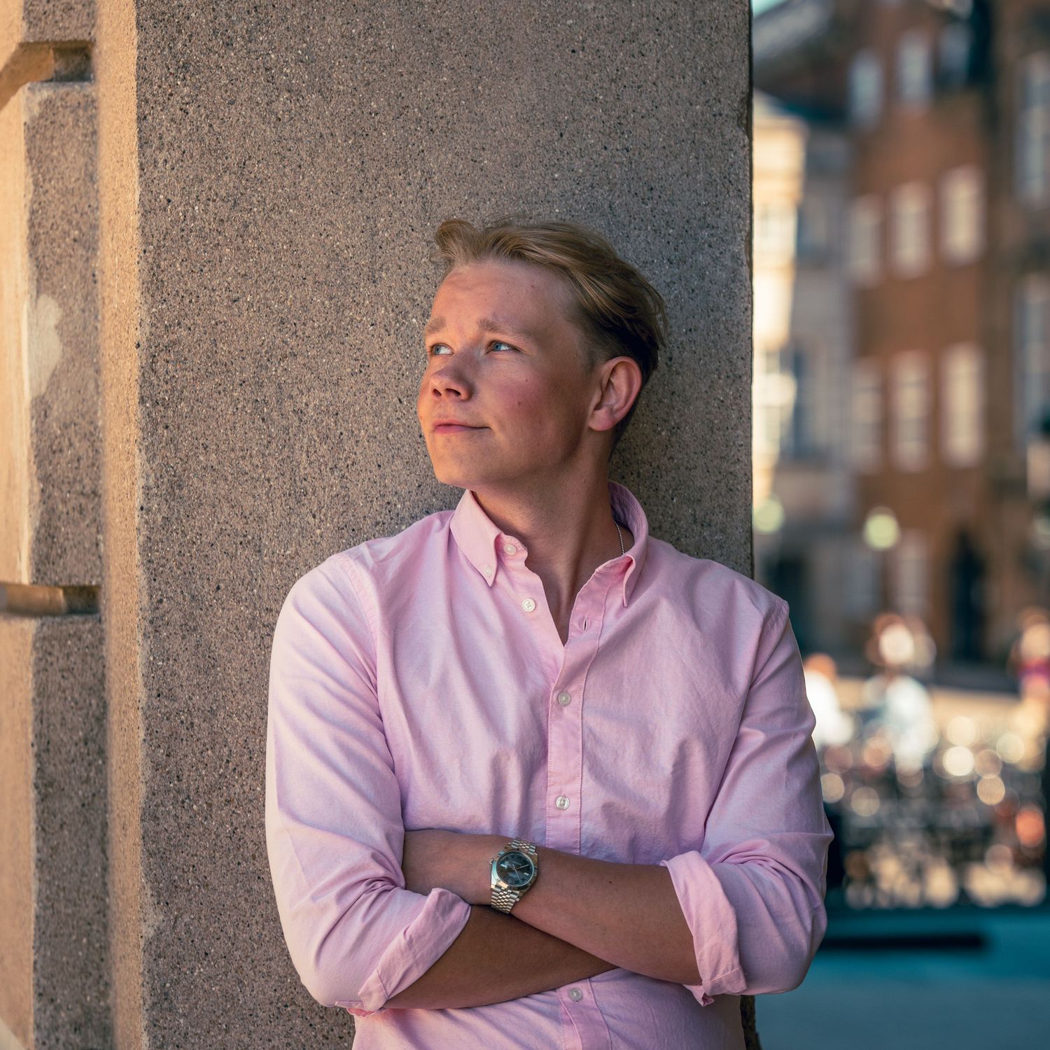 Person in a pink button-down shirt leans against a stone column with arms crossed in warm evening city light.