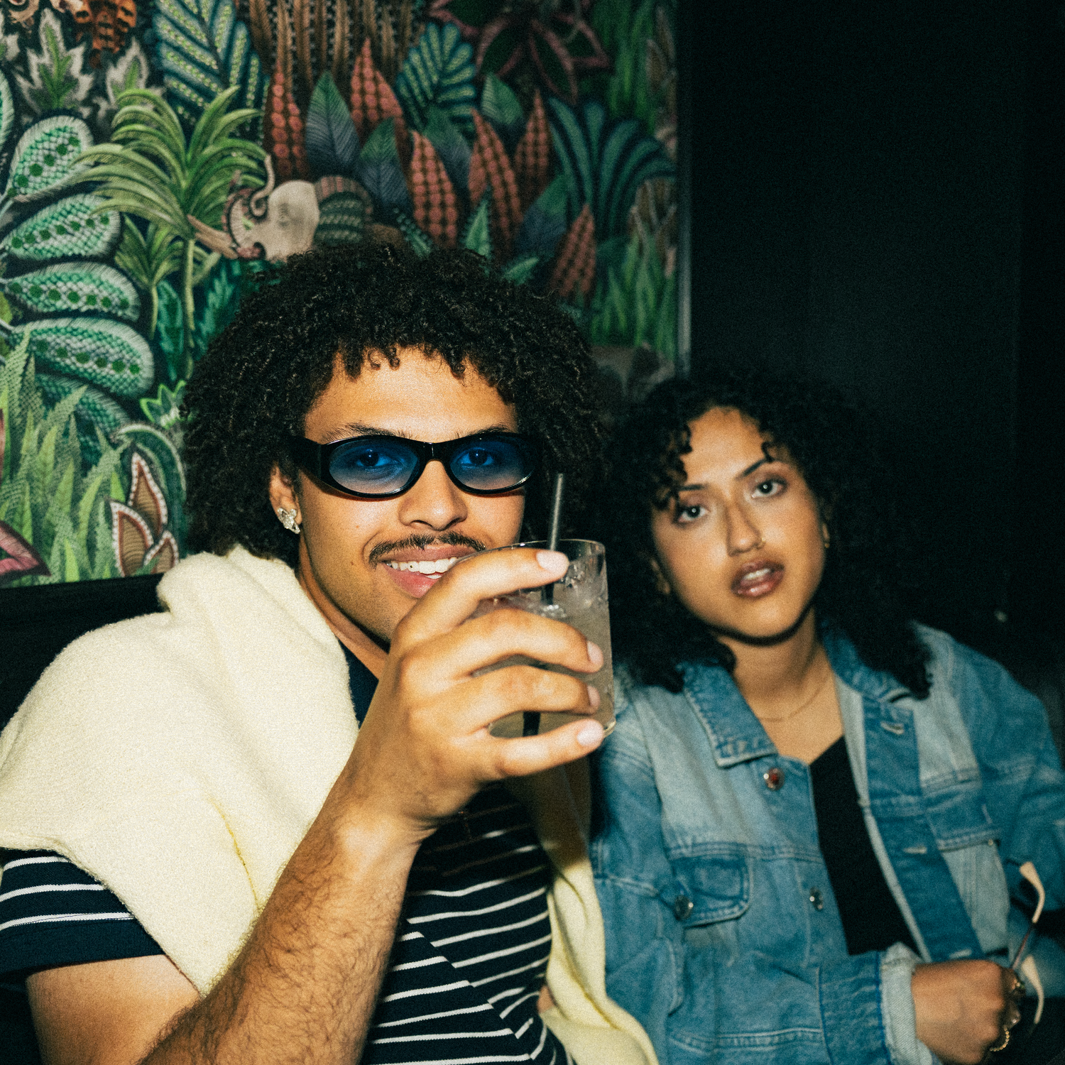 Two people enjoying drinks at a tropical-themed venue with lush green plant mural in the background.