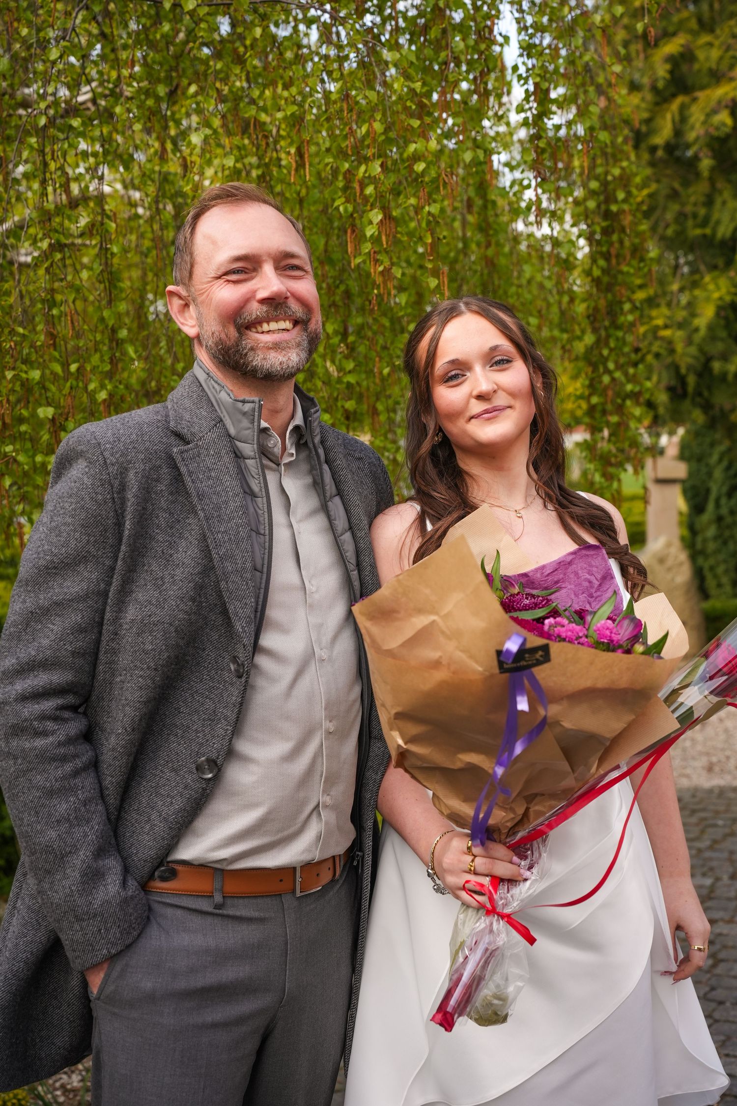 Two people in formal attire pose together outdoors with a bouquet of purple flowers and springtime trees in background.