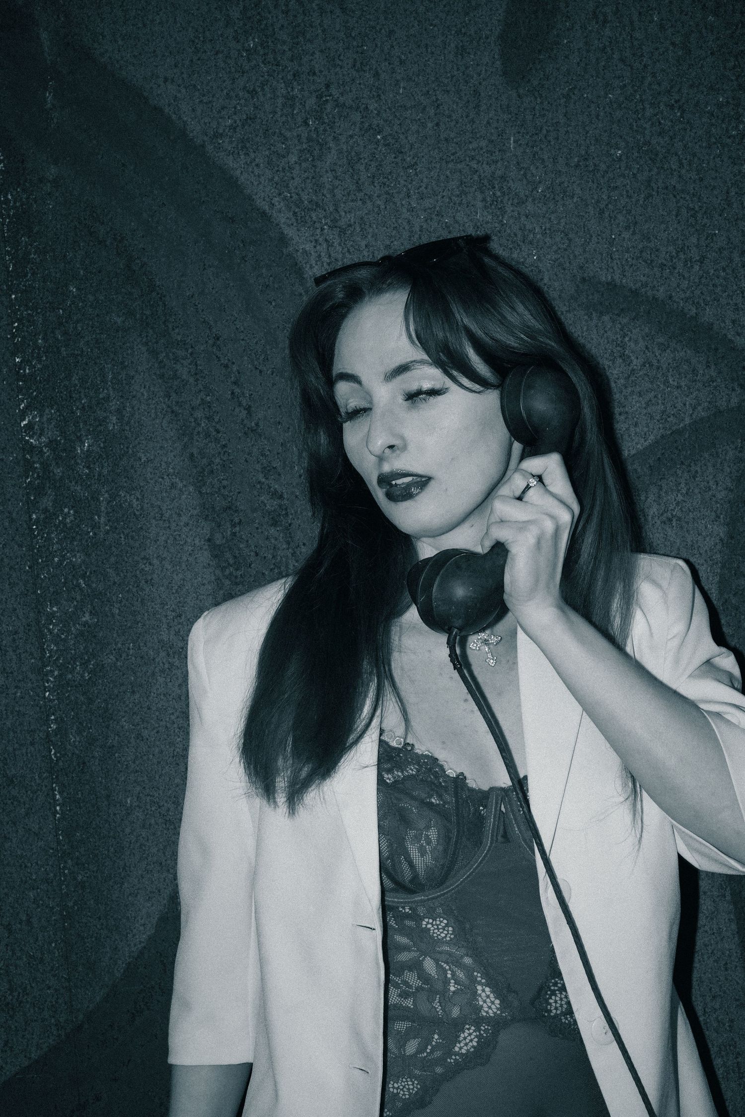 A person in a white blazer and lace top poses with a vintage telephone handset against a dark background.