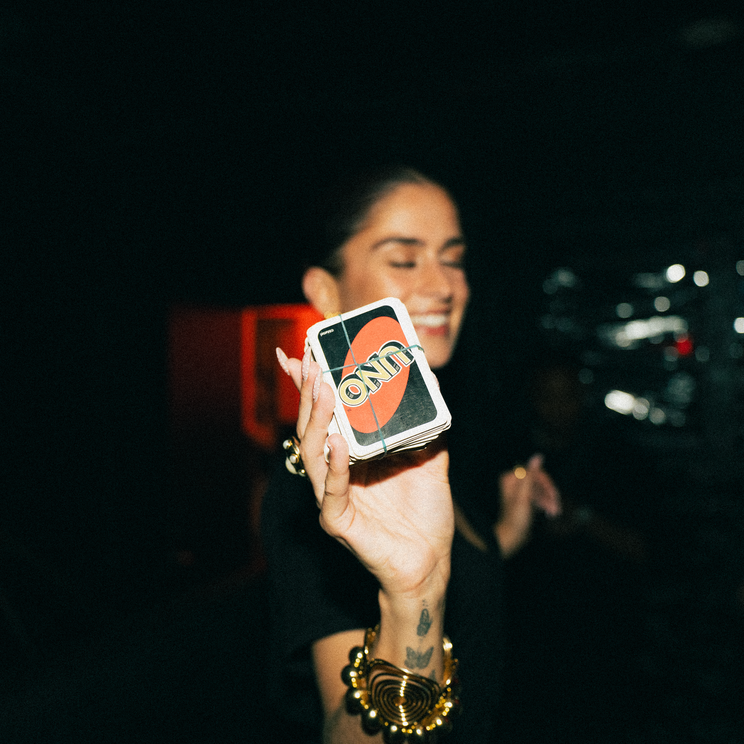 Hand holding a UNO card game in a dark nighttime setting with gold bracelet jewelry visible against blurred background.