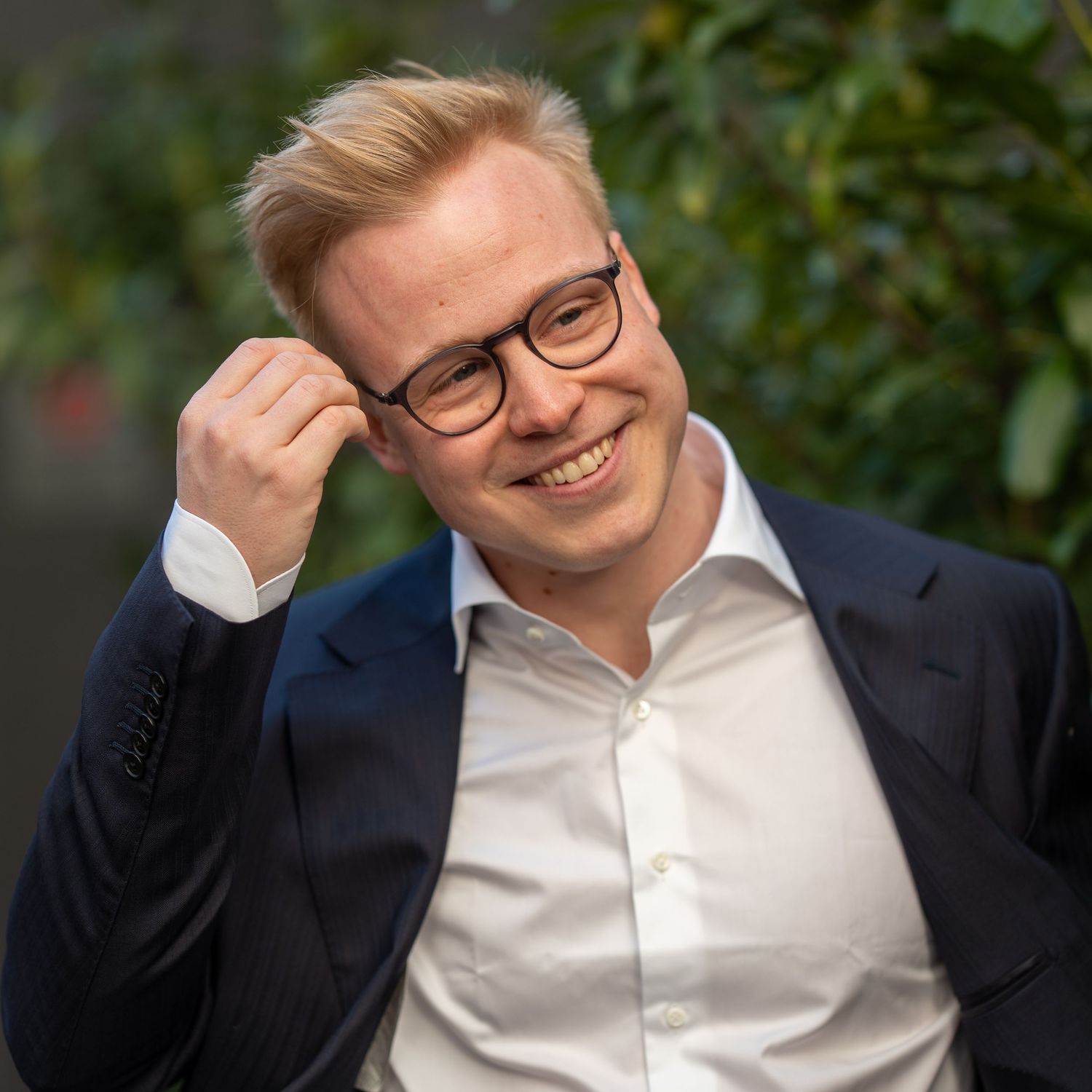 Professional headshot of person adjusting glasses while wearing suit and white shirt.