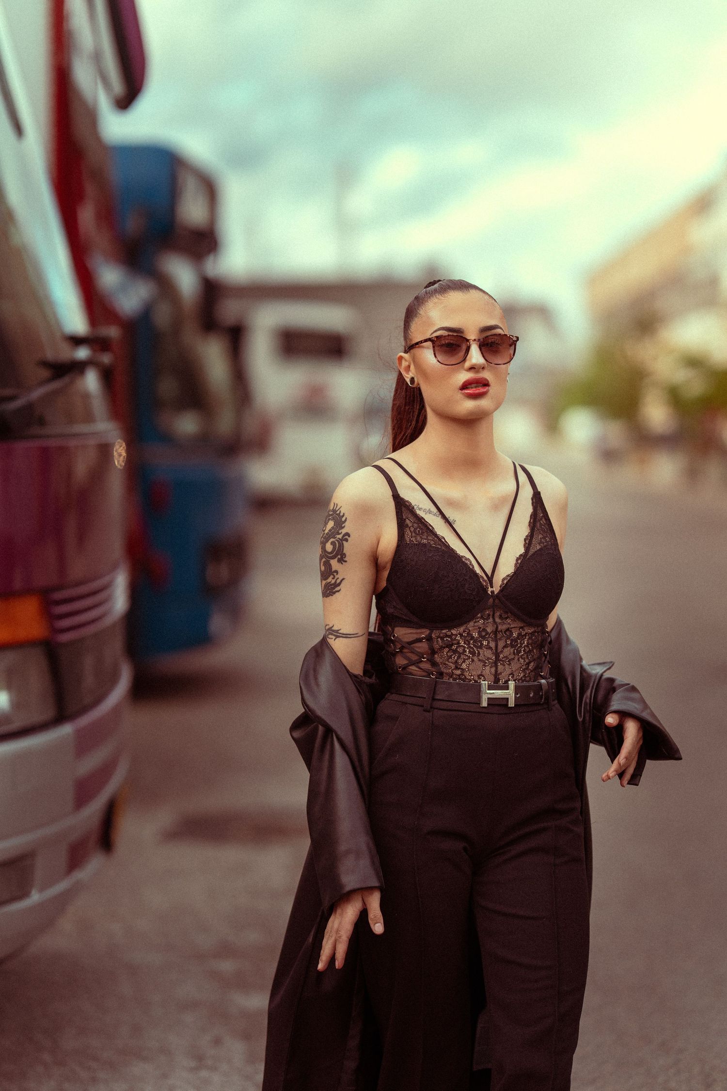 Fashionable person in sunglasses and dark outfit poses confidently between buses in an industrial setting at dusk.