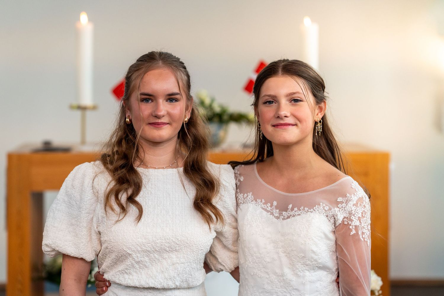 Two friends in white lace dresses pose together at a formal event with lit candles glowing in the background.