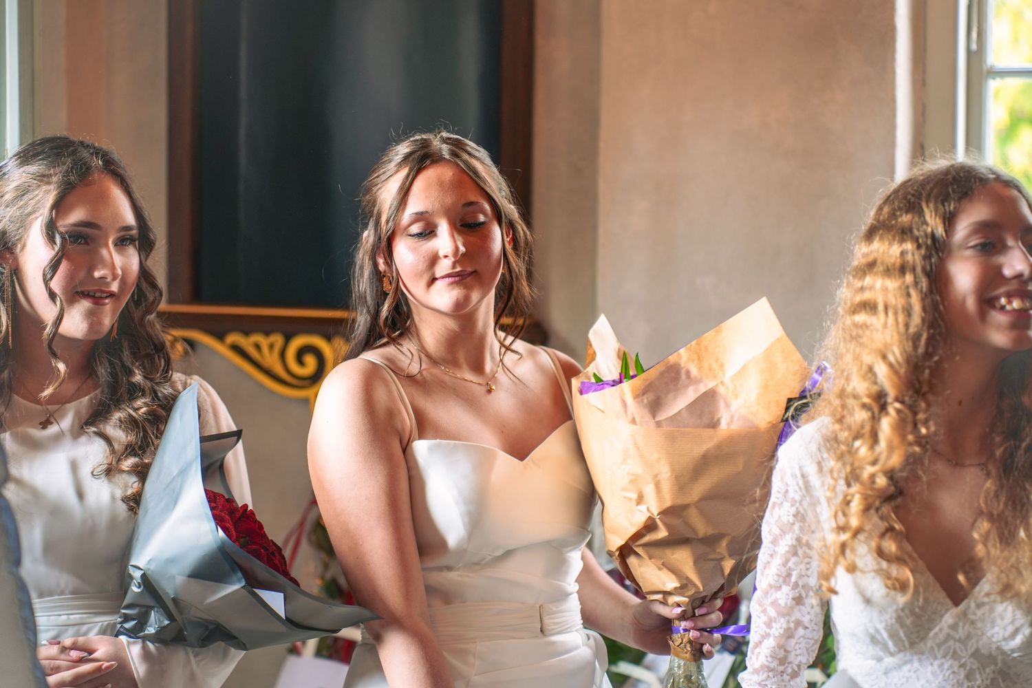 Three brides sitting together at a wedding reception share joyful laughter while holding floral bouquets.