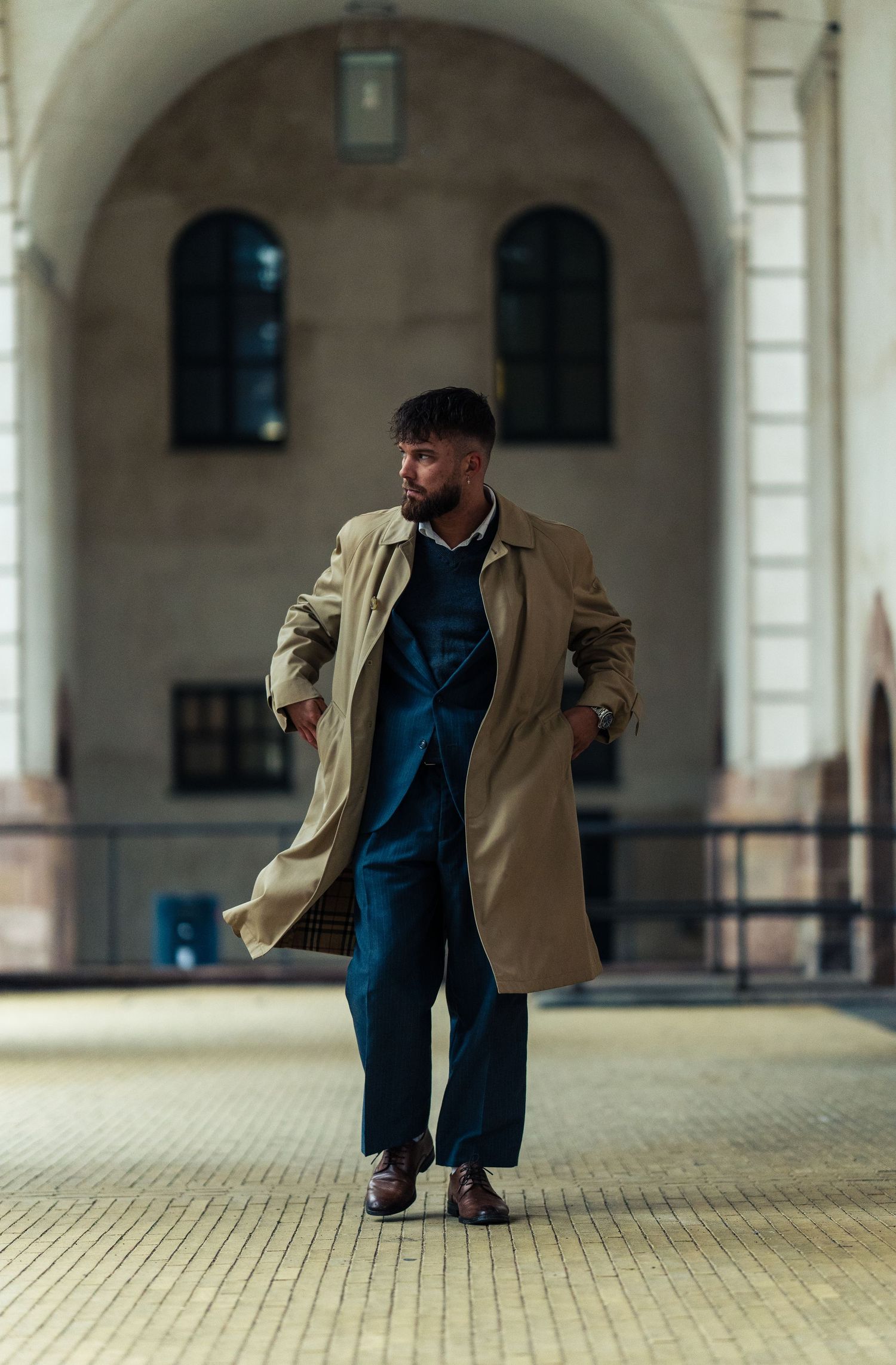 Man in beige trenchcoat and navy suit stands under arched passageway.