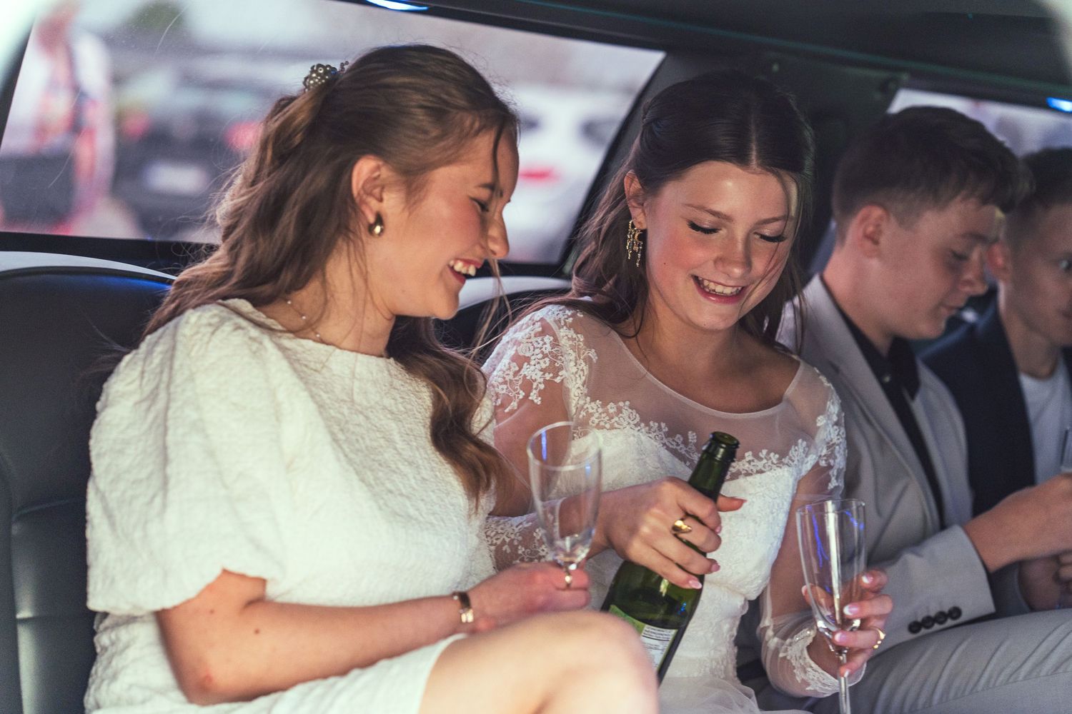 Two women in formal attire share drinks and laugh together while sitting in the backseat of a car.